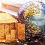 A miniature globe next to a miniature shipping container filled with cardboard shipping boxes sitting on a large keyboard.