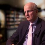 A man in a black suit and a light colored tie wearing glasses in front of an out of focus library discussing the value of a company wide perspective.