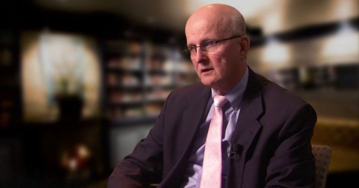 A man in a black suit and a light colored tie wearing glasses in front of an out of focus library discussing the value of a company wide perspective.