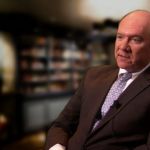 A man in a dark suit and blue checkered tie sitting down talking into a camera about potential reasons for GM's bankruptcy in front of an out of focus library.