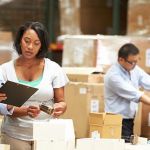 Purchasing manager working in a warehouse reviewing inventory sheets on a clipboard another employee is holding out for her to read, while a third employee works on taping up boxes to ship out.