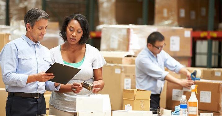 Purchasing manager working in a warehouse reviewing inventory sheets on a clipboard another employee is holding out for her to read, while a third employee works on taping up boxes to ship out.