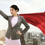 A woman in business attire and a red superhero cape pointing her finger in the air with the backdrop of a city behind her.