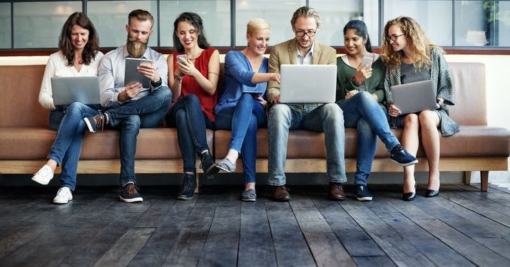 A group of seven people sitting on a couch together looking at different devices, such as laptops, tablets and smartphones, to depict how blended courses can be taken anywhere on any device.