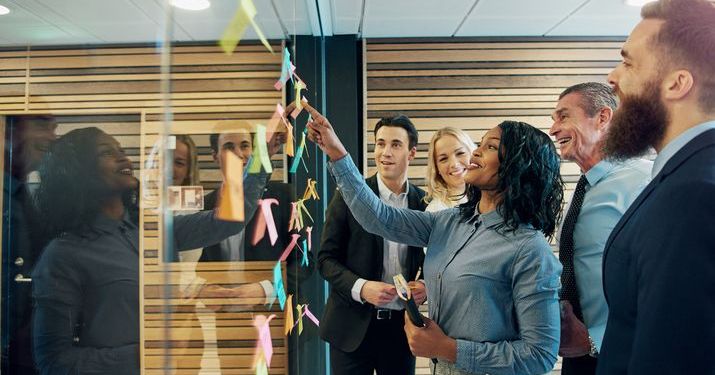 Group of employees working together in an office developing SMART goals by brainstorming and putting post it notes up on a glass.