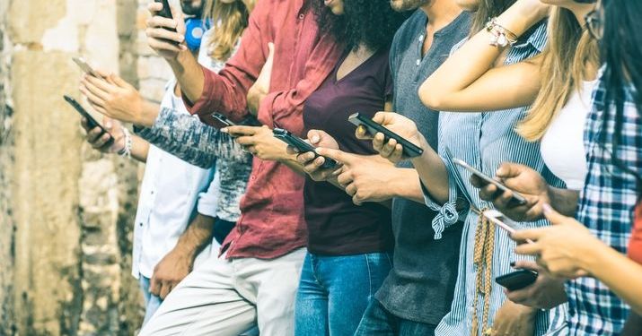 Millennials leaning against a wall using smartphones.