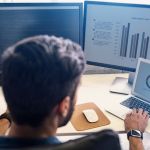 Employee sitting at his desk with two computer monitors and a laptop depicting various sets of business analytics data in the form of bat charts and graphics.