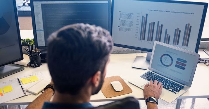 Employee sitting at his desk with two computer monitors and a laptop depicting various sets of business analytics data in the form of bat charts and graphics.