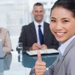 Four employees sitting at a conference room table developing global business strategy, with one woman looking into the camera and giving a thumbs up.