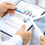 Close up of a pair of hands holding a clipboard with documents showing different types of charts, while another employee is holding a tablet next to him.