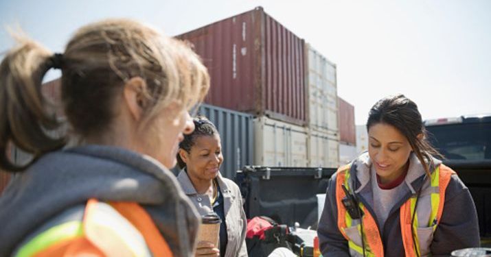 A group of women wearing reflective work vests in a shipyard surrounded by metal shipping containers, working on optimizing their supply chain.