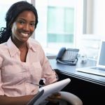 Woman holding a notepad and smiling at her desk in an office, with a laptop on the desk in front of her.