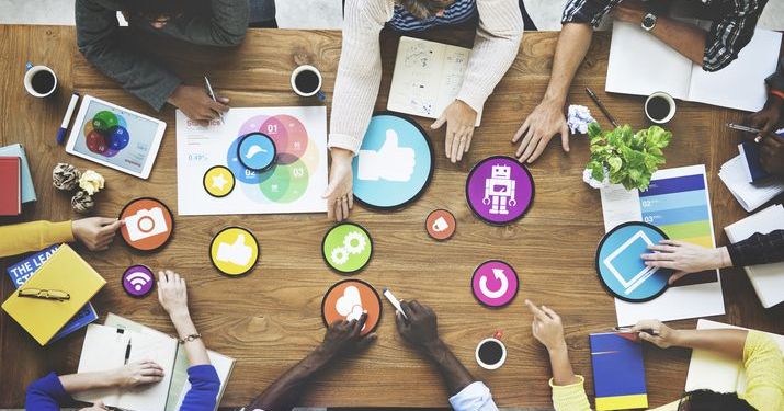 Group of employees working on brand journalism marketing around a table that has multiple printed graphics spread across it, representing different aspects of social media and digital marketing.