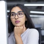 Woman wearing glasses looking a desktop computer screen trying to decide between an MBA and MHA.