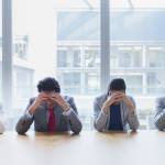 Four office employees in business clothes sitting in a row at table with both hands on their heads, feeling stressed out.