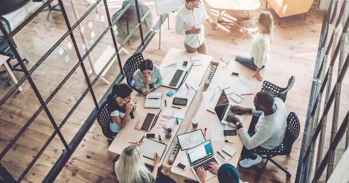 An overhead shot of a conference room with a successful team of eight employees working together on their laptops at a messy table.