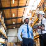 Two men wearing business clothes and white hard hats talking about supply chain in a warehouse filled with boxes of inventory.