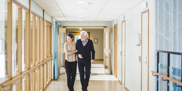 A young woman walking down the hall with an elderly man while holding his arm for support.