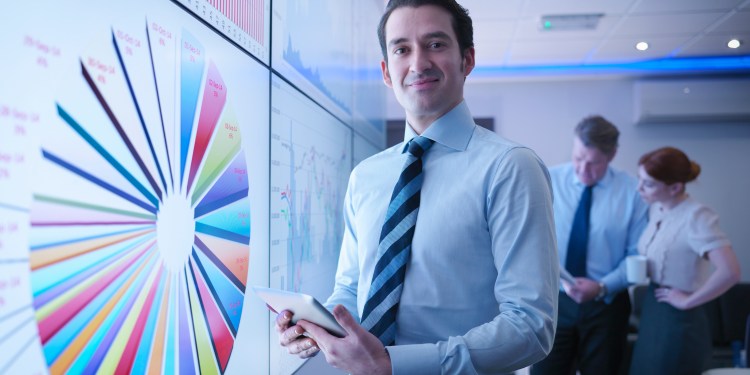 Male manager in front of data graphs on screen in meeting room with team members behind him analyzing data.