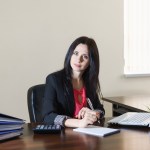 Female administrative services manager in a business suit taking notes on notepad at desk with stack of binders next to her.