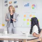 Female executive gesturing to white board while giving presentation to fellow professionals in office/workplace.