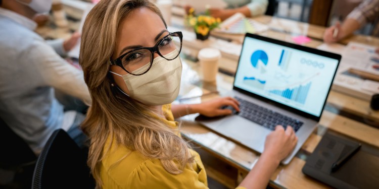 Woman looking at camera, wearing a facemask while working at the office. Data analytics and charts on her computer screen. She sits a table with co-workers, also masked.