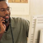 A man working on his computer while talking on the phone with a calendar on the wall behind him.