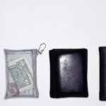 Four wallets lined up next to each other, three of them are dark leather and the fourth is clear plastic with money inside of it.