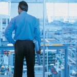 A man looking down at a warehouse from a large glass window.