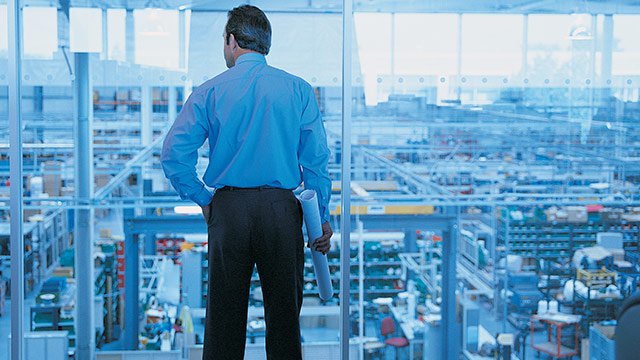 A man looking down at a warehouse from a large glass window.