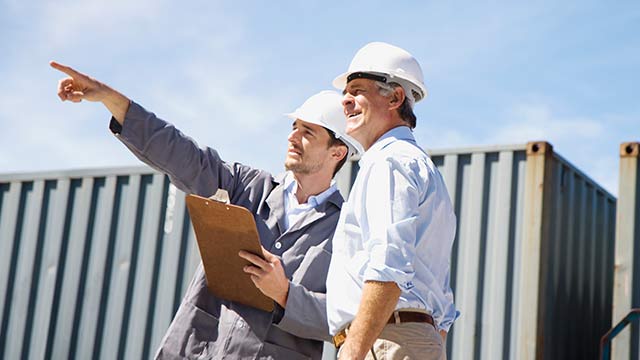 Two men in a shipping yard with hard hats on and one of them is pointing in the air while holding a clipboard.