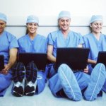 Four medical professionals wearing scrubs while sitting on the floor with their backs leaning up against the wall working on laptops.