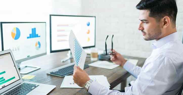 Business analyst analyzing business performance data while holding paper at desk