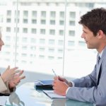 A businessman and a businesswoman sitting down across a table talking to each other.