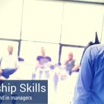 Leadership skills for managers depicted by co-workers sitting around a conference room table, listening to a man who is standing up and speaking about leadership skills managers should have.