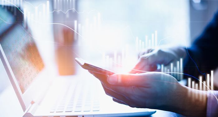 A pair of hands using a smartphone over a laptop that has holographic line charts floating around it.