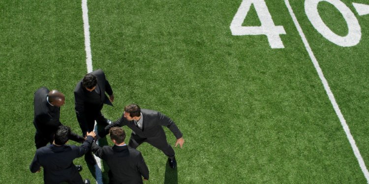 A team of businessmen in suits and ties on a football field in a huddle to represent teamwork.