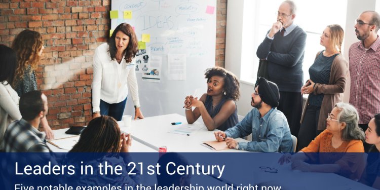 An office full of people working together talking to each other around a table with a large whiteboard on the wall that says "Ideas" in marker.