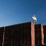 A man wearing a white coat and a hard hat standing on top of a tall stack of wooden pallets.