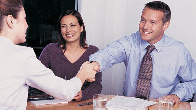 A man and a woman sitting at table meeting with another womam, introducing themselves and shaking hands.