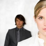 A close up headshot of a business woman wearing white, with a businessman wearing black stands against a white wall behind her.