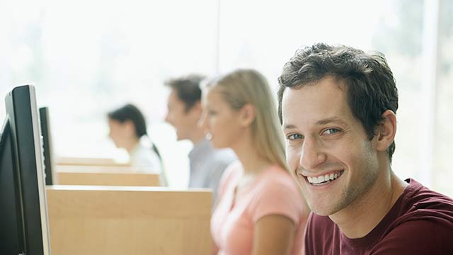 Four students sitting in a row in a computer lab with the man closest to the camera smiling.