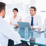 Three people sitting on one side of a glass table interviewing a man sitting across from them.