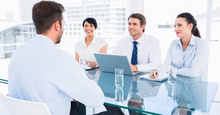 Three people sitting on one side of a glass table interviewing a man sitting across from them.