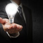 Close up of a man in a suit and tie holding out his hand with a light bulb floating in his hand.