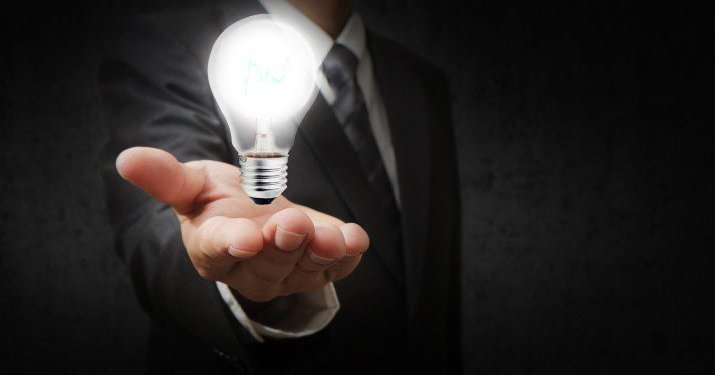 Close up of a man in a suit and tie holding out his hand with a light bulb floating in his hand.