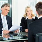 Three business people sitting around a table, with one man on one side of a table and a man and a woman sitting across from him looking at a piece of paper and talking to him as if they are interviewing him.