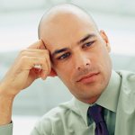 A man wearing a dress shirt and tie with his hand on his head looking down thinking.