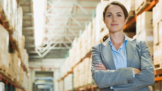 A woman in a warehouse with her arms crossed smiling into a camera.