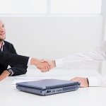 Two men shaking hands across a white table with a blue laptop on it.